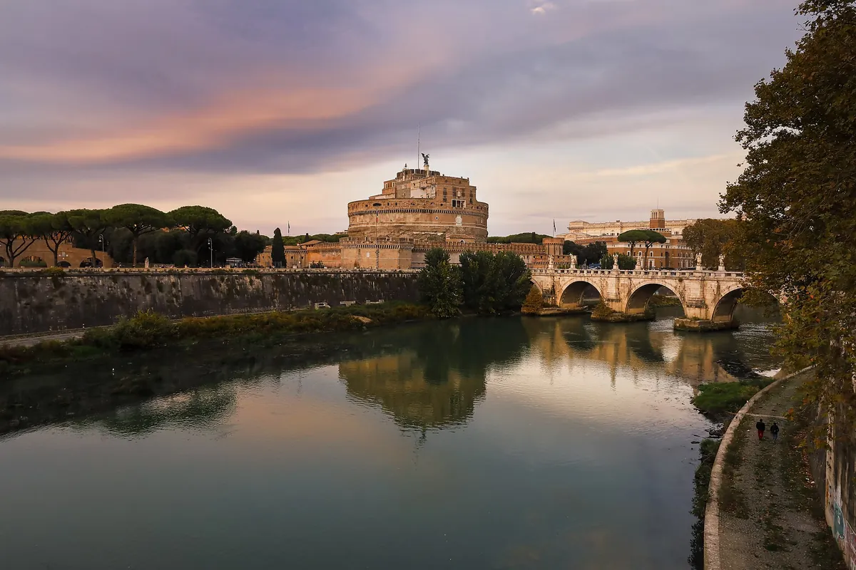 Castel Sant'Angelo a jeho most přes Tiberu při západu slunce, s růžovými mraky nad obzorem a kulatou pevností odrážející se v řece; třetí jednání Toscy se odehrává v jejích horních patrech.