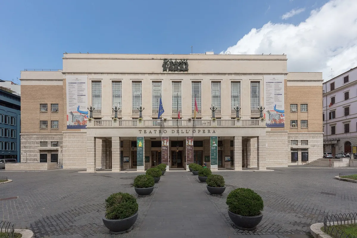 Světlé kamenné průčelí Teatro dell'Opera di Roma s balkonem, vysokými okny a názvem velkými písmeny nad vchodem.